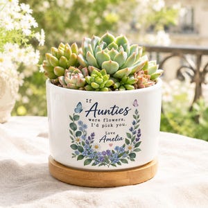 Op de afbeelding: Een witte keramische plantenpot met een verzameling groene vetplanten. De pot is voorzien van een bloemenkransontwerp met de tekst: If Aunties were flowers, I'd pick you. Love, Amelia. Het staat op een ronde lichte houten voet.