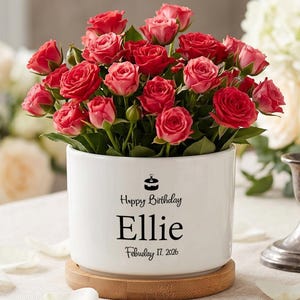 May include: A white ceramic planter filled with red and pink roses. The planter has the text "Happy Birthday Ellie February 17, 2026" in black. The planter sits on a wooden base.