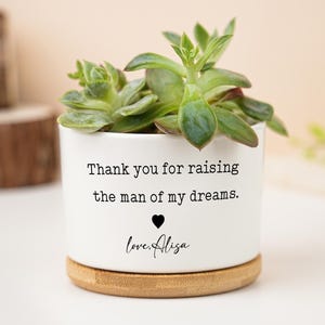 May include: A white ceramic planter with a succulent plant. The planter has a bamboo base and features the text "Thank you for raising the man of my dreams." with a heart and the name "love, Alisa."