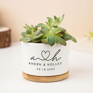 Op de afbeelding: Een witte keramische plantenbak met een vetplant. De plantenbak heeft een bamboe basis en heeft een zwart hartontwerp met de namen "Ahern & Holley" en de datum "03.18.2024".