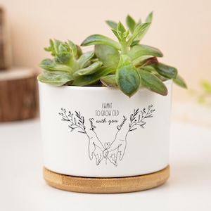 Puede incluir: Una maceta de cerámica blanca con una planta suculenta. La maceta tiene una base de bambú y presenta un dibujo lineal negro de dos manos que se sostienen, con el texto "I want to grow old with you" y los nombres "Emily" y "Daniel".