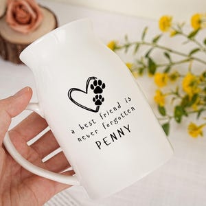 May include: White ceramic mug with a curved handle, featuring a black heart with paw prints and the text "a best friend is never forgotten PENNY". The mug has a wide opening and a smooth, glossy finish.