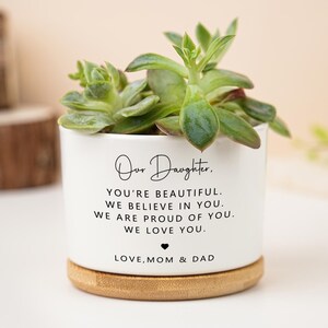 Może przedstawiać: Biała ceramiczna doniczka z sukulentem. Doniczka ma bambusową podstawę i zawiera tekst "Our Daughter. YOU'RE BEAUTIFUL. WE BELIEVE IN YOU. WE ARE PROUD OF YOU. WE LOVE YOU. LOVE, MOM & DAD."