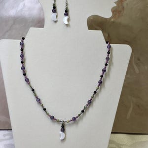 Può includere: Un set di collana e orecchini con un ciondolo a forma di mezzaluna e orecchini abbinati. Il set di gioielli include perline viola, nere e verdi. Il set è esposto su un espositore per gioielli bianco.