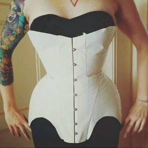 May include: A white corset with a black top and a row of buttons down the front. The corset is made of a sturdy fabric and has a fitted design.