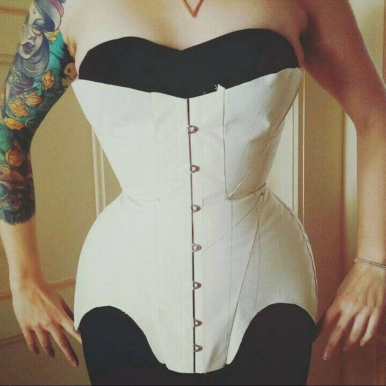 May include: A white corset with a black top and a row of buttons down the front. The corset is made of a sturdy fabric and has a fitted design.