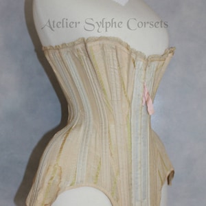 May include: A cream-colored corset with a striped pattern and a pink ribbon at the back. The corset is on a white mannequin and has the text "Atelier Sylphe Corsets" printed on the front.