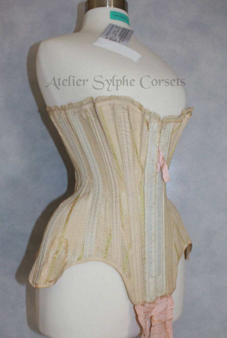 May include: A cream-colored corset with a striped pattern and a pink ribbon at the back. The corset is on a white mannequin and has the text "Atelier Sylphe Corsets" printed on the front.