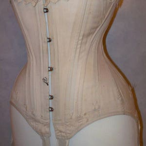 May include: A white cotton corset with a lace top and a row of metal hooks down the front. The corset has a long, pointed shape and is designed to cinch the waist. It has four white straps hanging from the bottom.