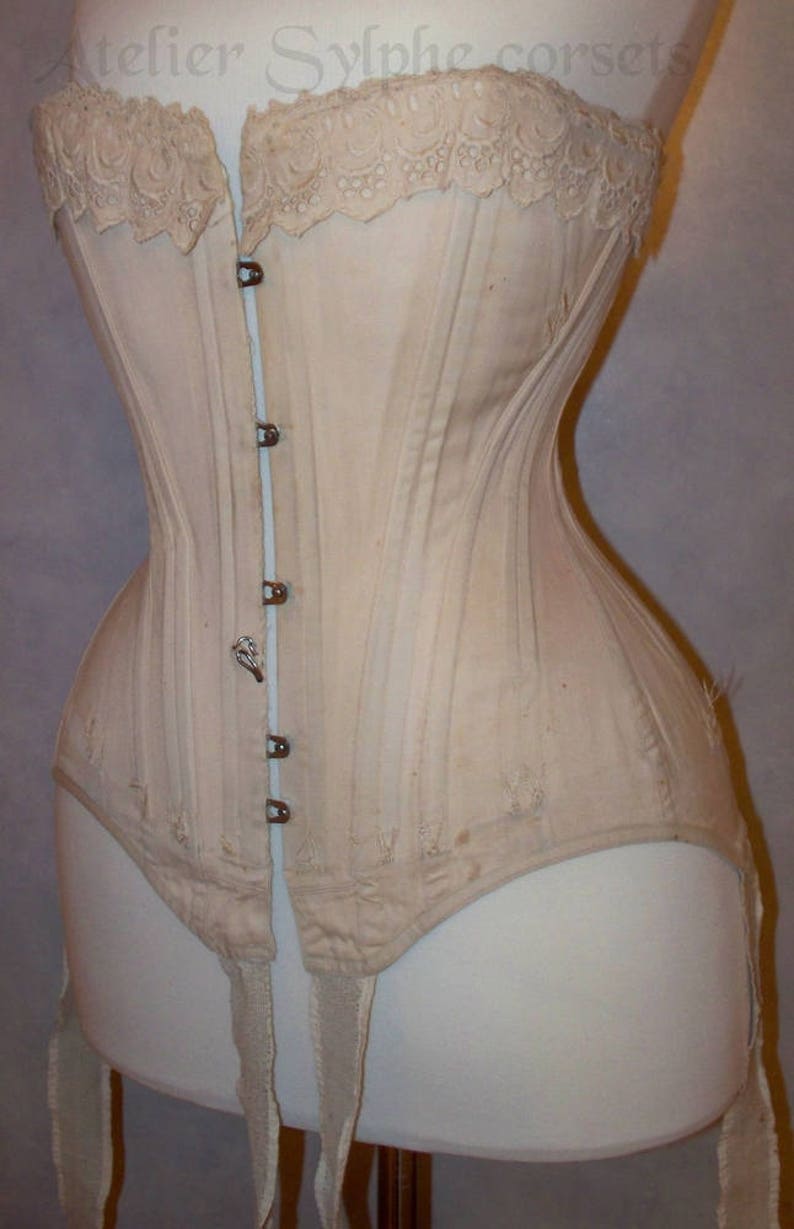 May include: A white cotton corset with a lace top and a row of metal hooks down the front. The corset has a long, pointed shape and is designed to cinch the waist. It has four white straps hanging from the bottom.