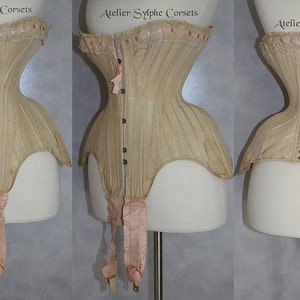May include: A beige corset with a lace trim and pink ribbon accents. The corset is laced up the back with black laces. The corset is on a white mannequin.