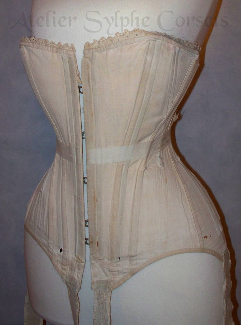 May include: A white cotton corset with a lace trim and a long busk closure. The corset has a series of vertical panels and is designed to cinch the waist. The corset is displayed on a white dress form.
