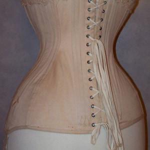 May include: A beige corset with a lace trim and a long white lacing in the back. The corset is on a white dress form. The text "Atelier Solrhie corsets" is visible at the top of the image.