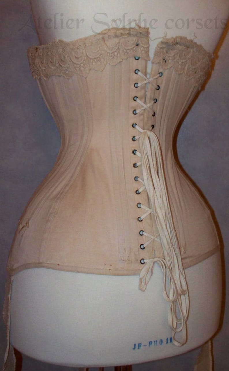 May include: A beige corset with a lace trim and a long white lacing in the back. The corset is on a white dress form. The text "Atelier Solrhie corsets" is visible at the top of the image.