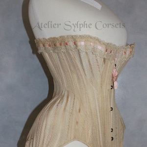 May include: A beige corset with pink ribbon trim and lace detailing. The corset is on a white dress form and has a label that reads "Atelier Sylphe Corsets".