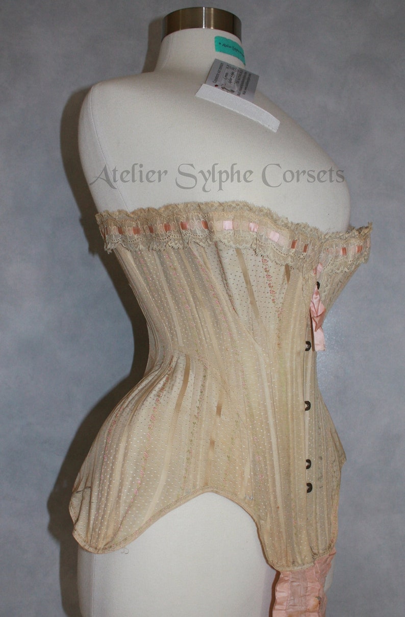 May include: A beige corset with pink ribbon trim and lace detailing. The corset is on a white dress form and has a label that reads "Atelier Sylphe Corsets".
