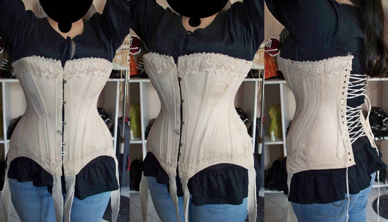 May include: A white cotton corset with lace trim and a front closure. The corset has a boned structure and is shown being worn by a person.