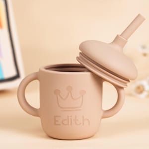 K&ouml;nnte beinhalten: Ein hellbrauner Silikon-Trinkbecher mit einem Kronenmuster und dem eingravierten Namen "Edith" auf der Vorderseite. Der Becher hat zwei Griffe und einen passenden Deckel mit Strohhalm. Der Becher ist f&uuml;r Babys und Kleinkinder konzipiert.