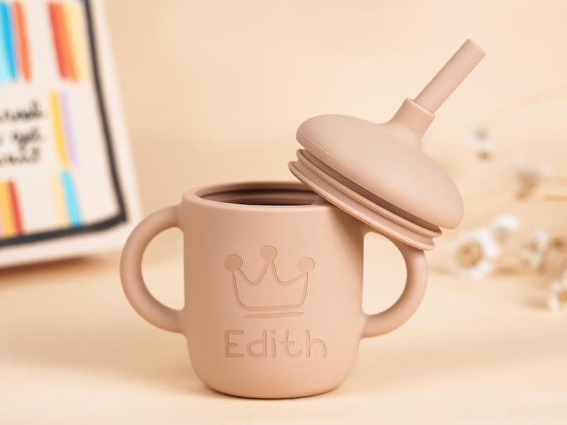 K&ouml;nnte beinhalten: Ein hellbrauner Silikon-Trinkbecher mit einem Kronenmuster und dem eingravierten Namen "Edith" auf der Vorderseite. Der Becher hat zwei Griffe und einen passenden Deckel mit Strohhalm. Der Becher ist f&uuml;r Babys und Kleinkinder konzipiert.
