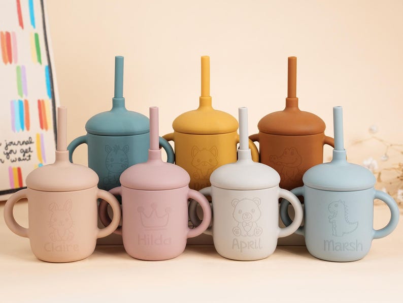 K&ouml;nnte beinhalten: Eine Sammlung farbenfroher Silikon-Trinkbecher mit Griffen und Strohhalmen. Jeder Becher hat eine andere Pastellfarbe mit passendem Deckel und Strohhalm. Die Becher sind mit Tierdesigns und Namen verziert.