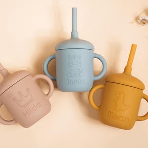 K&ouml;nnte beinhalten: Drei Babybecher aus Silikon in Pastellfarben: Rosa, Blau und Gelb. Jeder Becher hat einen Strohhalm, Griffe und einen Deckel. Der rosa Becher hat eine Krone und den Namen "Hilda". Der blaue Becher hat ein Zebra und den Namen "Jonas". Der gelbe Becher hat einen Dinosaurier und den Namen "Marsh".