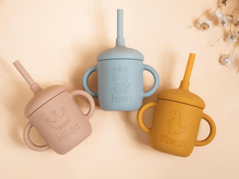 K&ouml;nnte beinhalten: Drei Babybecher aus Silikon in Pastellfarben: Rosa, Blau und Gelb. Jeder Becher hat einen Strohhalm, Griffe und einen Deckel. Der rosa Becher hat eine Krone und den Namen "Hilda". Der blaue Becher hat ein Zebra und den Namen "Jonas". Der gelbe Becher hat einen Dinosaurier und den Namen "Marsh".