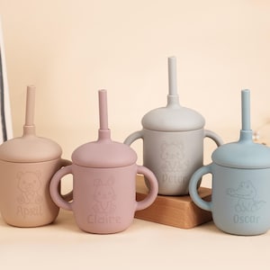 K&ouml;nnte beinhalten: Vier Babybecher aus Silikon in Pastellfarben: Beige, Rosa, Grau und Blau. Jeder Becher hat einen Deckel mit Strohhalm und zwei Griffen. Die Becher sind mit Tierillustrationen und Namen verziert: April, Claire, Peter und Oscar.