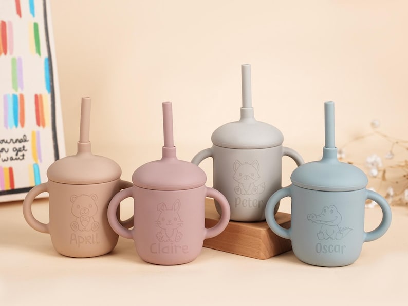 K&ouml;nnte beinhalten: Vier Babybecher aus Silikon in Pastellfarben: Beige, Rosa, Grau und Blau. Jeder Becher hat einen Deckel mit Strohhalm und zwei Griffen. Die Becher sind mit Tierillustrationen und Namen verziert: April, Claire, Peter und Oscar.