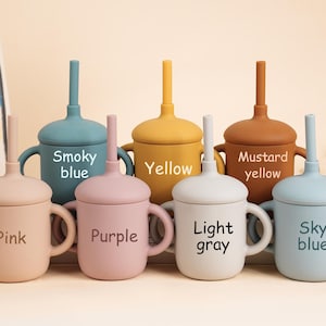 K&ouml;nnte beinhalten: Eine Sammlung farbenfroher Babybecher aus Silikon mit Deckel und Strohhalm. Die Becher sind in den Farbt&ouml;nen Rosa, Lila, Hellgrau, Himmelblau, Rauchblau, Gelb und Senfgelb gehalten. Jeder Becher hat einen Griff und den Farbnamen aufgedruckt.