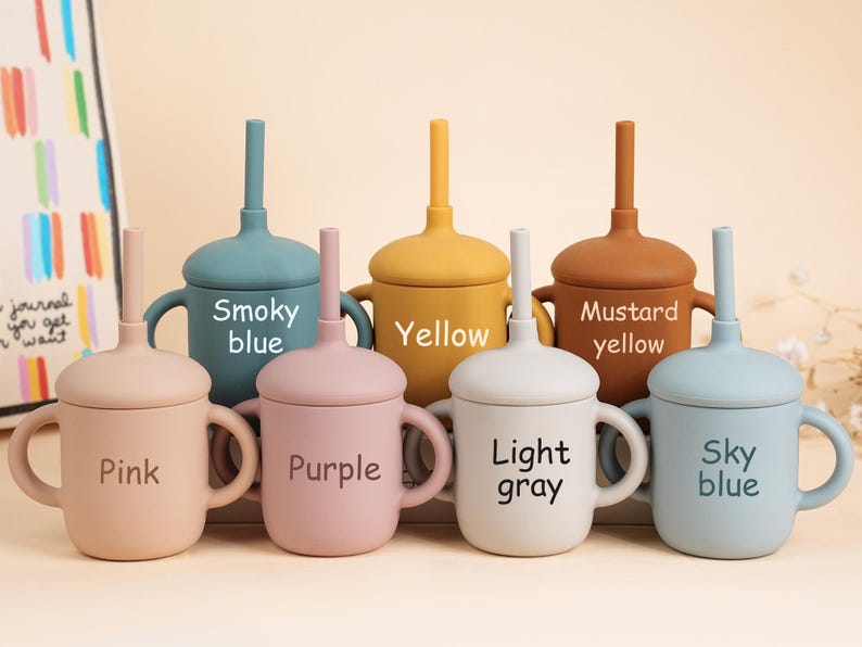K&ouml;nnte beinhalten: Eine Sammlung farbenfroher Babybecher aus Silikon mit Deckel und Strohhalm. Die Becher sind in den Farbt&ouml;nen Rosa, Lila, Hellgrau, Himmelblau, Rauchblau, Gelb und Senfgelb gehalten. Jeder Becher hat einen Griff und den Farbnamen aufgedruckt.
