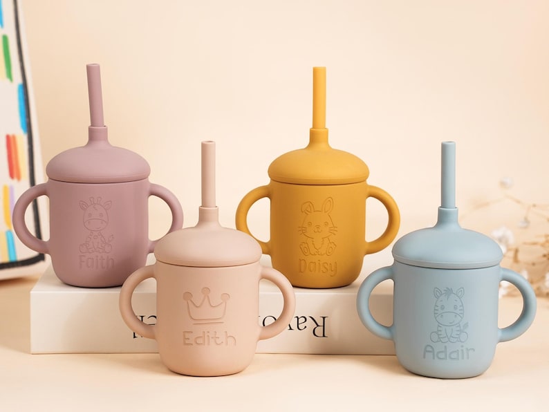 K&ouml;nnte beinhalten: Vier Babybecher aus Silikon mit Deckeln und Strohhalmen in ged&auml;mpften Farben: Mauve, Pfirsich, Senfgelb und Blau. Jeder Becher hat ein anderes Tierdesign und einen Namen. Die Becher haben Griffe.