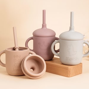 K&ouml;nnte beinhalten: Eine Sammlung von Babybechern aus Silikon in verschiedenen Rosa- und Beiget&ouml;nen. Jeder Becher hat einen Deckel und einen Strohhalm. Ein Becher hat ein Zebra-Design und den Namen "Jonas", ein anderer eine Krone und den Namen "Hilda", und der letzte einen Dinosaurier und den Namen "Marsh".