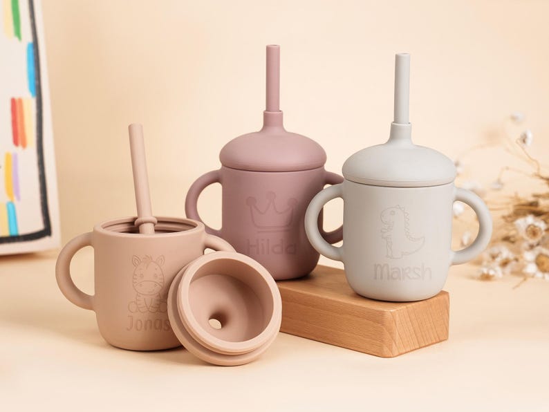 K&ouml;nnte beinhalten: Eine Sammlung von Babybechern aus Silikon in verschiedenen Rosa- und Beiget&ouml;nen. Jeder Becher hat einen Deckel und einen Strohhalm. Ein Becher hat ein Zebra-Design und den Namen "Jonas", ein anderer eine Krone und den Namen "Hilda", und der letzte einen Dinosaurier und den Namen "Marsh".