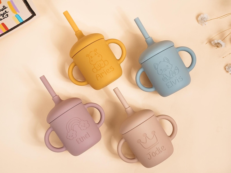 K&ouml;nnte beinhalten: Vier Silikon-Trinkbecher in Gelb, Blau, Mauve und Beige. Jeder Becher hat einen passenden Strohhalm und Griffe. Die Becher sind mit gepr&auml;gten Designs eines Nilpferds, eines Fuchses, eines Regenbogens und einer Krone mit den Namen Aries, Elvis, Ethel und Jodie verziert.