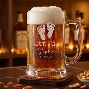 Op de afbeelding: Een heldere bierpul gevuld met bier en schuim. De pul heeft een handvat en is gegraveerd met babyvoetjes, de tekst "HAPPY 1ST FATHER'S DAY", de naam "Sophia" en de datum 15/06/2026. De pul staat op een houten onderzetter.