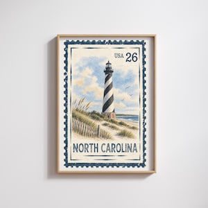 May include: A framed print designed as a postage stamp featuring a black and white spiral-striped lighthouse on a sandy beach with dunes and grass. The text reads USA 26 and NORTH CAROLINA in a dark blue font against a light sky background.