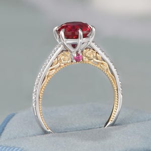 May include: A ring featuring a round, deep red center stone held by silver prongs. The band is two-toned, with gold floral engravings on the sides and silver edges lined with small clear stones. A tiny red accent stone sits below the main setting.