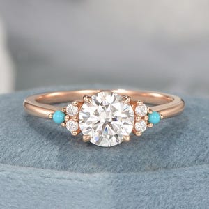 May include: Rose gold ring featuring a large, round, clear gemstone as the centerpiece. The ring is accented with small, clear gemstones and turquoise-colored stones on either side of the center stone. The ring is set on a blue surface.