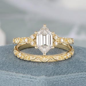 May include: A gold engagement ring set with a large, clear, hexagonal-cut gemstone. The band features intricate floral and leaf patterns, with smaller accent stones. The ring sits on a blue surface.