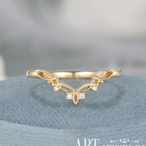 Peut inclure: Une bague dorée avec un design incurvé, ornée de petites pierres précieuses claires. La bague présente un motif délicat et orné avec un élément central en forme de fleur. La marque "APT JEWELRY" est visible en bas.