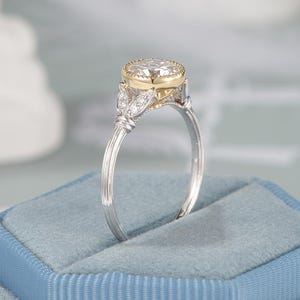 May include: A two-tone engagement ring with a large, round, clear gemstone set in a gold bezel. The ring features a silver band with intricate leaf-like details and small accent stones. The ring is displayed on a blue velvet ring box.