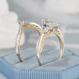 May include: Two engagement rings with intricate designs. One ring features a large, clear gemstone and smaller blue stones, set in a silver and gold band. The other ring has a similar design. Both rings are displayed on a blue velvet ring box.