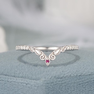 May include: A silver-toned ring with a V-shaped design. The ring features a small, round, red gemstone at the center of the V. The band is adorned with small, clear gemstones. The ring is set on a blue surface.