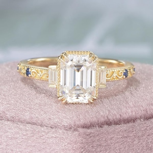 Puede incluir: Un anillo de oro con una piedra central transparente de talla esmeralda. El anillo tiene un diseño de filigrana detallado en la banda, con pequeñas piedras azules y una piedra rectangular a cada lado de la piedra principal.