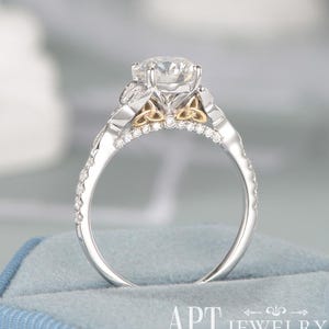 May include: A silver-toned ring featuring a large round center stone and a band accented with small pavé-set stones. The setting includes gold-toned Celtic knot details on the sides. The text APT JEWELRY is visible at the bottom of the image.
