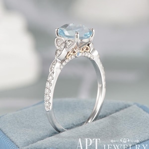 May include: A silver-toned ring featuring a large, oval-cut light blue gemstone set in a prong setting. The band and the gallery are adorned with small, clear pavé-set stones, with intricate metalwork details visible beneath the main stone.