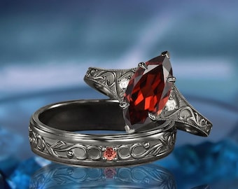 May include: A dark metal ring set featuring a marquise-cut red gemstone centerpiece and intricate scrollwork engravings. The band includes a small embedded red stone and clear accent stones, showcasing a detailed floral-inspired metal pattern.