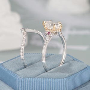 May include: Two rings displayed on a blue velvet surface. One ring features a large, rectangular clear stone with a gold setting and small pink stones. The other ring is silver with small clear stones.