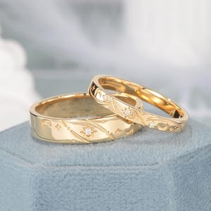 May include: Two gold bands featuring engraved celestial motifs including stars and crescent moons. The wider band displays a prominent star with a small embedded clear stone, while the thinner band features smaller star accents with clear stones.