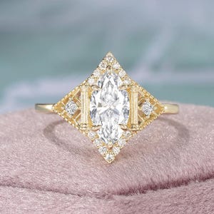 Puede incluir: Anillo de compromiso dorado con un diamante central de talla marquesa. El diamante está rodeado por un halo de diamantes más pequeños, engastados en un diseño geométrico. El anillo tiene una banda delicada y se muestra sobre una superficie rosa.